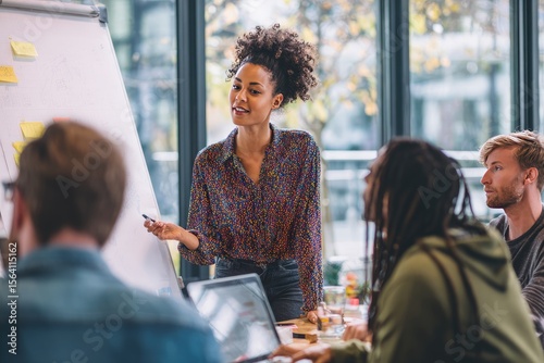 A young woman with curly hair leads a team meeting, presenting information on a whiteboard with sticky notes, surrounded by colleagues in a modern, bright office space