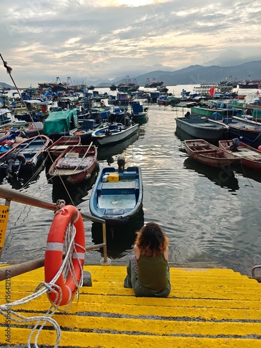 A woman relaxes on yellow steps near a harbor, observing numerous fishing boats bobbing on calm water during sunset. The sky is painted with soft clouds and colors. photo