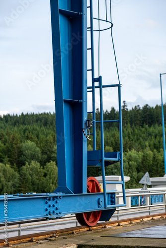 Close-up of a red wheel on a blue gantry crane moving along a rusty rail. Mechanical structure with bolts and ladder, surrounded by green forest in background.