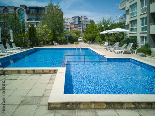 Clean outdoor pool with clear blue water, sun loungers, and umbrellas near modern apartment building. Peaceful summer vibe and relaxation area.