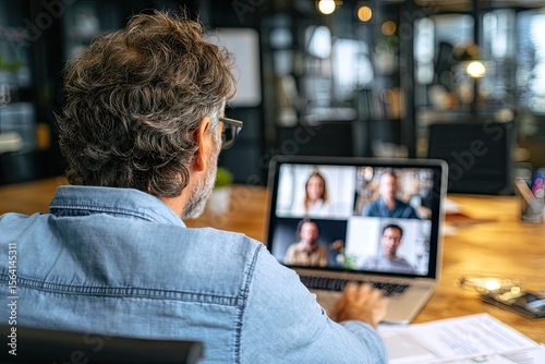 Rear view of man in denim shirt, glasses, participating in a video conference on laptop at a modern wooden desk