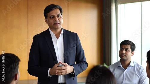 Man in a suit gesturing while addressing a group of people in a brightly lit office space area
