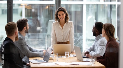 A smiling young businesswoman leads a diverse team meeting around a large table in a modern office, using laptops and documents