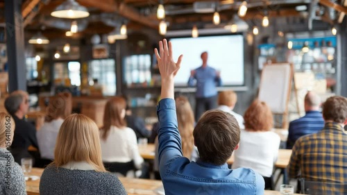 A male participant lifts his hand amid a bustling workshop as the presenter explains at the front. Concept of interactive learning fostering active engagement.