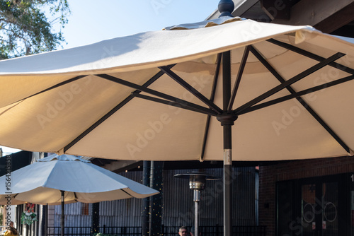 Multiple tan-colored fabric sunshades opened on a patio with black metal poles and hand cranks. The parasol has vents at the top of the poles. There are propane patio heaters among the beige shades.