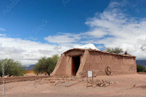 Main adobe chapel with sloping walls and a cartwheel, at El Mayorazgo Complex, Tinogasta, Catamarca. An icon of the Adobe Route under a blue sky with clouds.