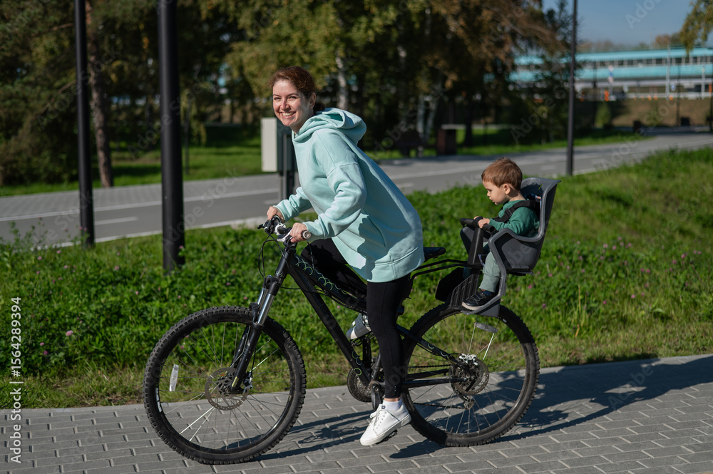 Fototapeta premium Caucasian woman riding bicycle with toddler son sitting behind her in child seat in park.