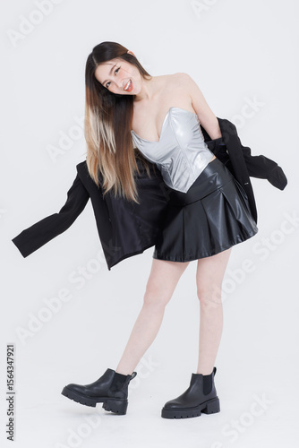 Fashion asian pretty young woman model pose playfully confident wearing silver corset top black pleated leather skirt black jacket expressing trendy streetwear style creative against white background.