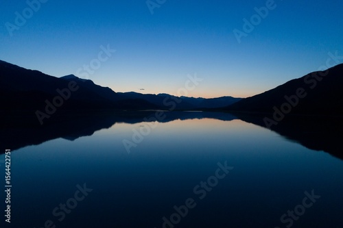Silhouetted peaks are casting reflection on lake water and dusky sky is fading in flat design