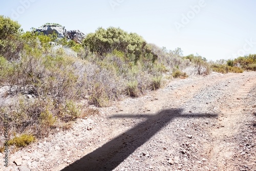 Shadow of cross is stretching across dusty unpaved road in flat vector design, with scrub bushes
