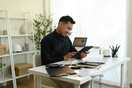 A Confident, Mature Asian businessman reviewing documents on a tablet while working in a modern, sunlit office