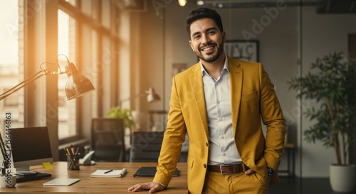 Man in yellow suit smiles at office desk with computer and bright window