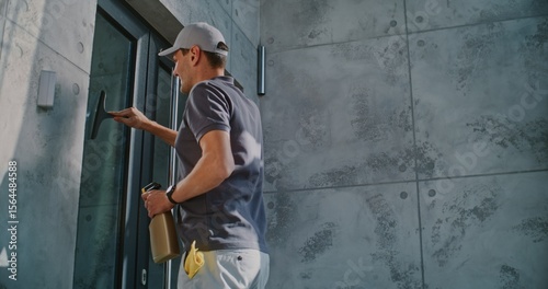 Professional Cleaner Spraying Cleaning Liquid and Washing Glass Door Outdoors of Modern Office Building. Businesswoman Going Out and Enters in Office. Male Specialist Working in Cleaning Service.