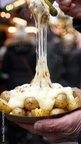 An extreme close-up video of warm-toned hands holding a paper plate of roasted potatoes with gooey raclette cheese, creating a long stretchy cheese pull from a pickled gherkin. Blurred festive