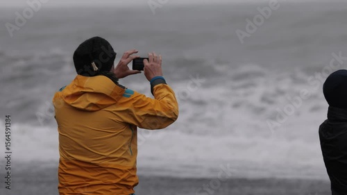 Person in a bright yellow raincoat taking a photo with a smartphone by the stormy sea, with waves crashing and another figure in the background, capturing a moody coastal scene.