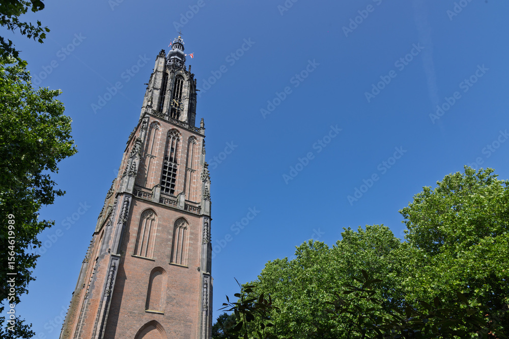 Fototapeta premium The imposing Onze Lieve Vrouwetoren of Amersfoort pierces a pristine blue sky, with lush green trees framing its ancient brick structure.