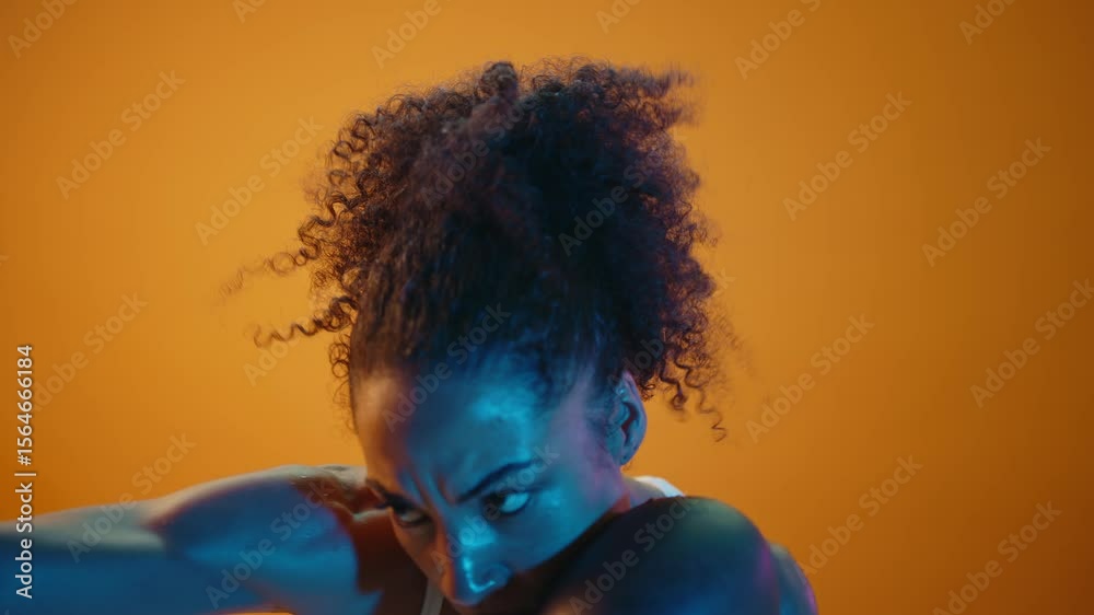 Female boxer wearing training gloves, throwing powerful punches against vibrant orange backdrop, illuminated by dramatic blue lighting during intense workout session