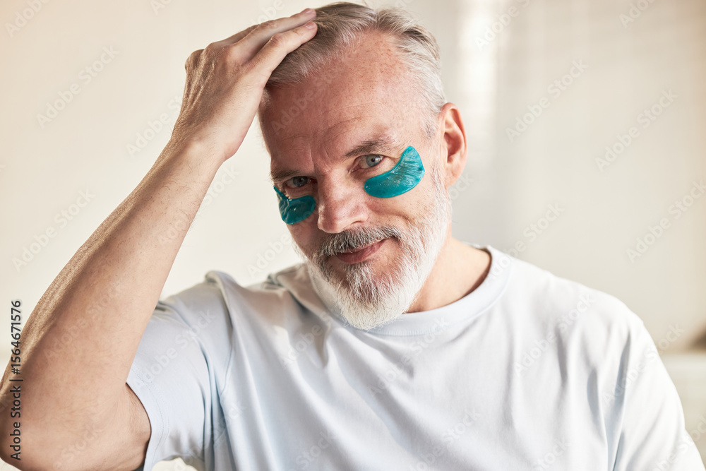 Obraz premium Portrait of middle aged Caucasian man smiling at camera, applying blue under eye patches, touching hair, demonstrating skincare routine in bright indoor setting