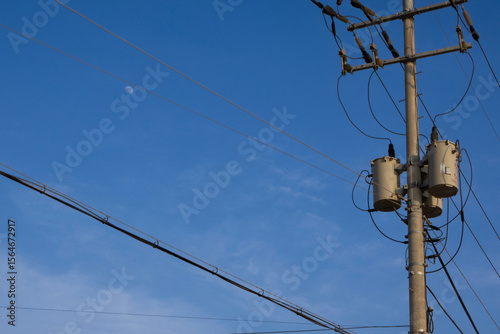 Electric poles on the street, transformers and wires hanging from them, and half a moon floating in the blue sky