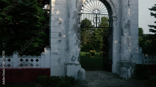 Aerial drone shot flying through a grand, old, weathered white gate with wrought iron details, revealing a beautiful, lush green garden with trees and manicured hedges.

