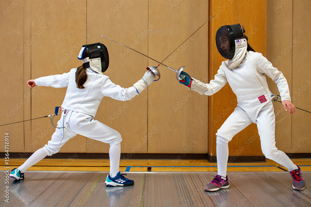Fototapeta premium Two fencers in white gear lunge toward each other during an intense bout on an indoor fencing piste.