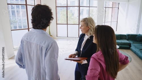 Woman in business attire presents a spacious apartment to a couple, highlighting large windows and urban views, showcasing potential living space
