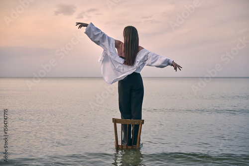Young woman stands gracefully on chair in shallow sea water, arms outstretched, capturing essence of freedom and balance against tranquil sunset backdrop