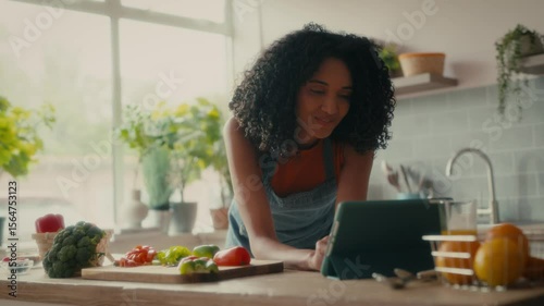 A woman with curly hair is preparing lunch in a bright, cosy kitchen. There are bowls, vegetables and a tablet on the counter.