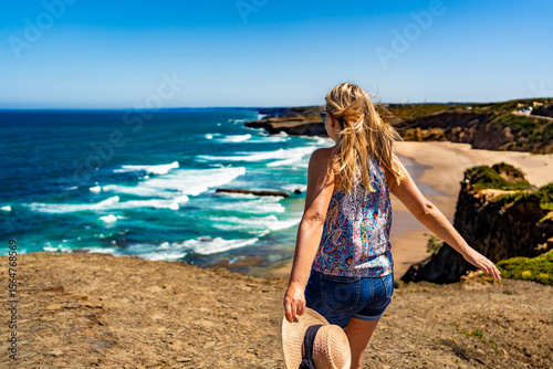 Beautiful mid adult woman tourist enjoying walk on cliff overlooking Monte Clerigo beach and ocean in summertime. Algarve Portugal. Back view