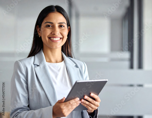 Portrait of young Hispanic professional business woman standing in office. Happy female company executive, smiling businesswoman entrepreneur corporate leader manager looking at camera using tablet.
