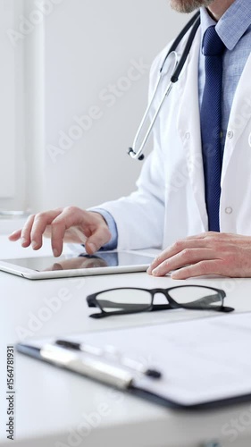 Male doctor wearing lab coat and stethoscope using digital tablet while sitting at desk in medical office, with medical chart and pen in the foreground