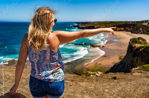 Beautiful mid adult woman tourist standing on cliff overlooking Monte Clerigo beach and ocean and pointing at something on sunny summer day. Algarve coast in Portugal. Back view