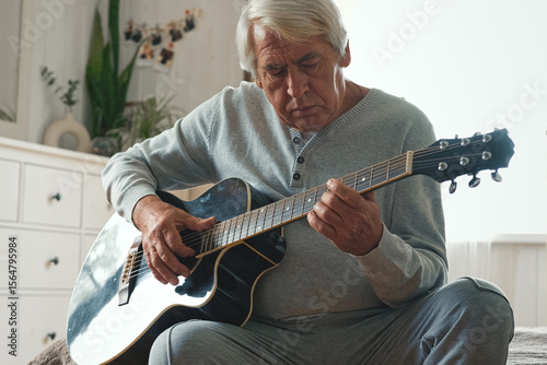 Senior man is playing guitar. Elderly male sitting at sofa and play music. Portrait of a gray haired mature 60s person with acoustic guitar, he is learning to play. Enjoying retirement life at home.