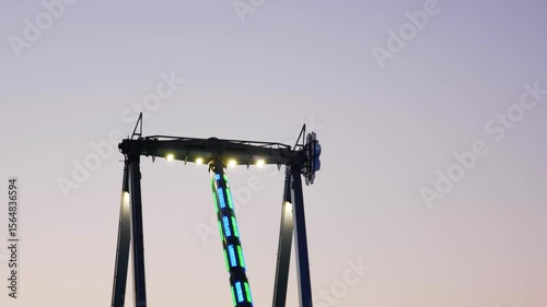 Wide shot of thrill seekers swinging back and forth on a giant Frisbee pendulum ride under a pastel sunset sky