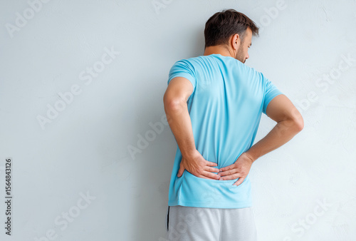 A man in dressed in a light blue tee and grey pajamas, stands against a white wall, clutching his back in pain from sciatica, his posture reflects sadness and physical discomfort.