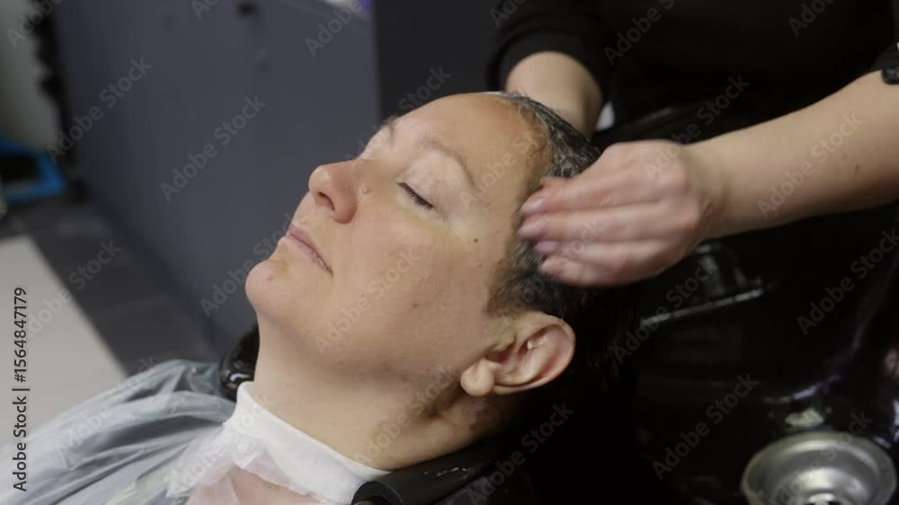 Professional hairdresser massaging shampoo into relaxed client's wet hair while leaning over salon sink, providing gentle hair care treatment with focused technique