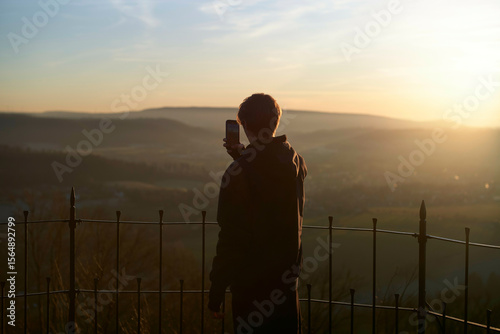 A person capturing the sunrise over rolling hills with a smartphone from behind a metal fence. Weser Uplands, Germany