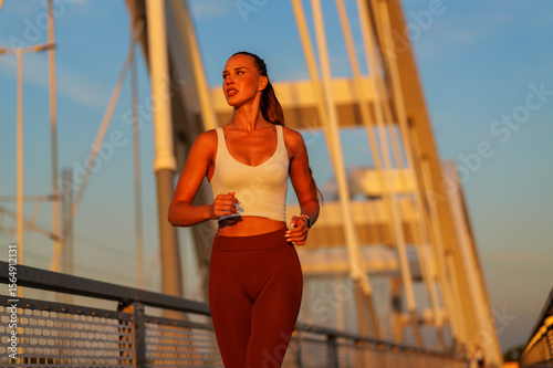 Young sporty woman running on a bridge at sunset, savoring her daily workout routine while embodying a commitment to a healthy lifestyle and fitness