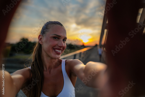 Sporty woman capturing a joyful selfie on a bridge during a stunning sunset after her workout, celebrating her commitment to a healthy lifestyle and fitness journey