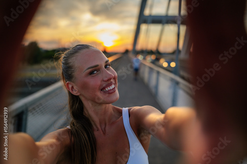 Smiling sportswoman taking a selfie on a bridge during sunset, enjoying the warm golden hour light after a refreshing workout, celebrating an active and healthy lifestyle