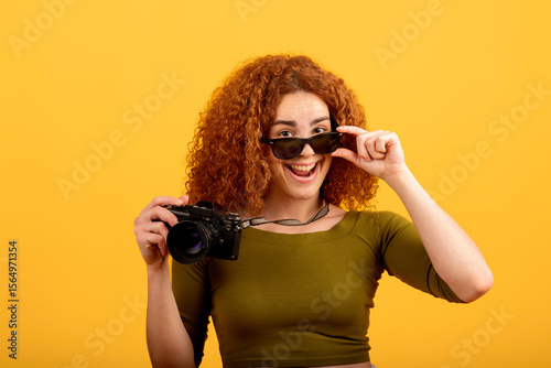 Tablou pe pânză Redhead photographer holding camera and lowering sunglasses on yellow background