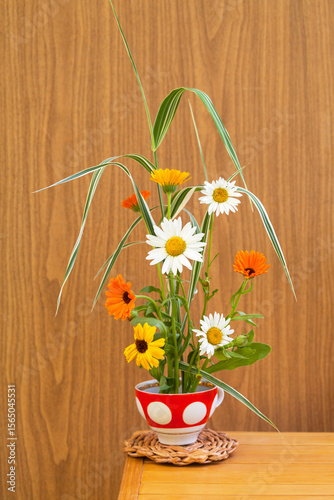 Beautiful rural floral arrangement of seasonal fresh garden flowers of marigolds and daisies on kenzan holder in cup in traditional ikebana on wooden wall background. Floral design for home as hobby