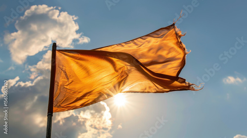Vibrant orange flag waves gracefully against backdrop of fluffy clouds and bright sun, creating striking contrast in sky. fabric billows gently, capturing light beautifully