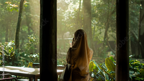 Woman in hijab gazes at sunlit tropical forest from indoors, a serene and contemplative moment with nature.