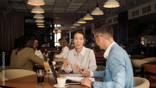 Asian woman and coworker talking about work while using laptop in modern coffee shop.
