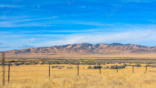 Barbed wire fence crossing dry grassland in Nevada desert landscape with mountains and blue sky