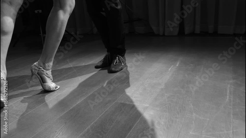 Black and white slow motion close-up of the legs and feet of a man and woman dancing a passionate Argentine tango on a wooden floor under dramatic spotlights.
