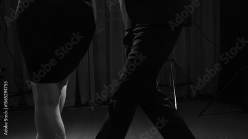 Black and white close-up on the legs and feet of a man and woman dancing a passionate Argentine tango, with dramatic shadows on a wooden floor.
