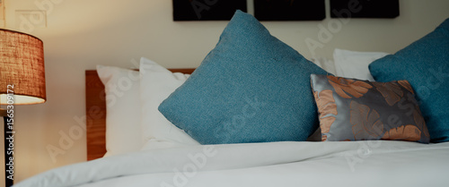 Close-up of pillows arranged on a neatly made bed with wooden lamp and headboard. Soft textures and neutral tones create a calm bedroom scene.