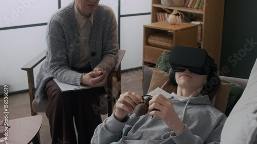 Medium shot of teenage patient lying on couch in VR headset touching fake spider to overcome arachnophobia during individual therapy session with unrecognizable psychologist